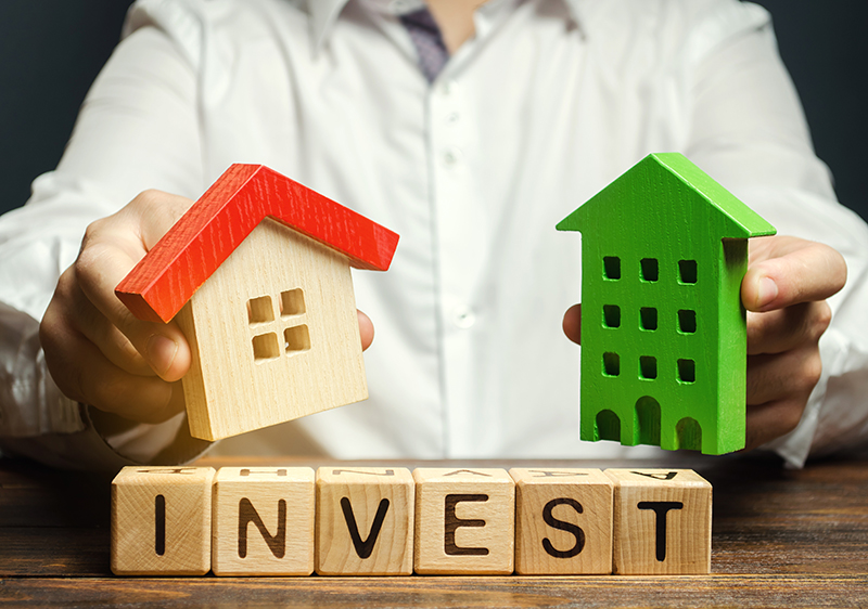 Wooden blocks with the word Invest, and houses in the hands of a businessman. The concept of investing in real estate construction. Effective investment. Buying apartments. Business and finance - JN Bank