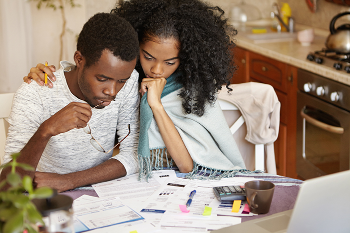 Young African-American couple facing financial problem. Unemployed male holding glasses, looking at papers on table with frustrated expression, not able to pay off utility bills, his wife hugging him - JN Bank