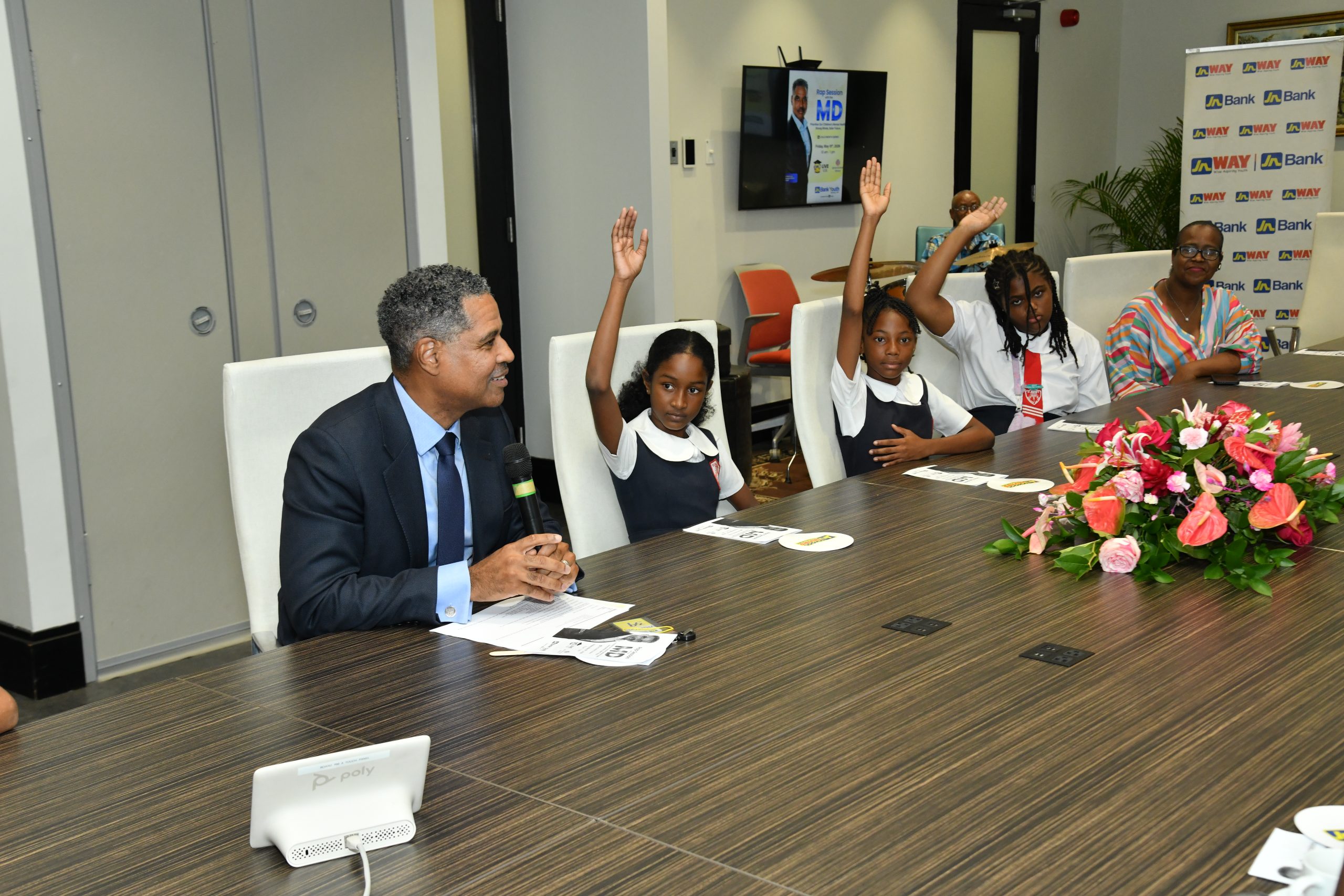 MD Rap Session - JN Bank Man in a suit speaks into a microphone at a conference table as four young girls raise their hands in a classroom-style panel, with a flower arrangement in the foreground and banners in the background.