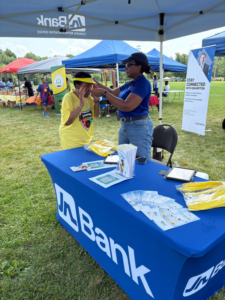 20260808_175507000_iOS - JN Bank Two women at a Brampton outreach booth under blue canopies at an outdoor community fair; a volunteer in a blue shirt helps a woman in a yellow shirt with a headband, standing beside a table covered with a blue cloth and brochures.
