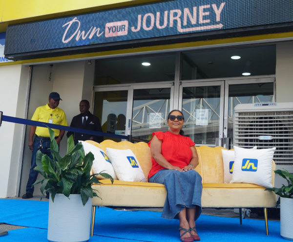 DSC03622 - JN Bank Woman in red blouse and sunglasses sits on a yellow sofa during a ribbon-cutting outside a store with an 'Own Your Journey' sign above the entrance.